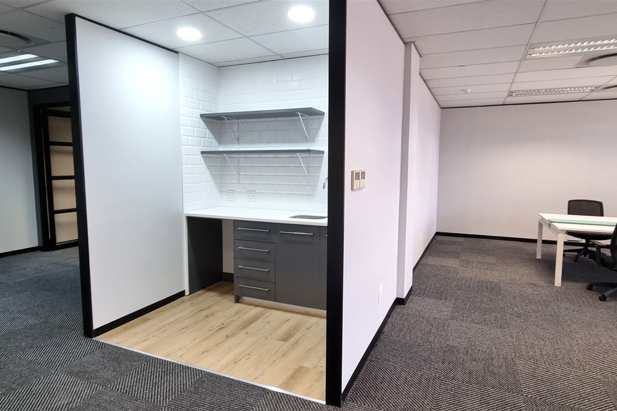 A modern office kitchenette area featuring built-in cabinetry, open shelving, and a tiled backsplash, integrated into an open-plan workspace with carpeted flooring.