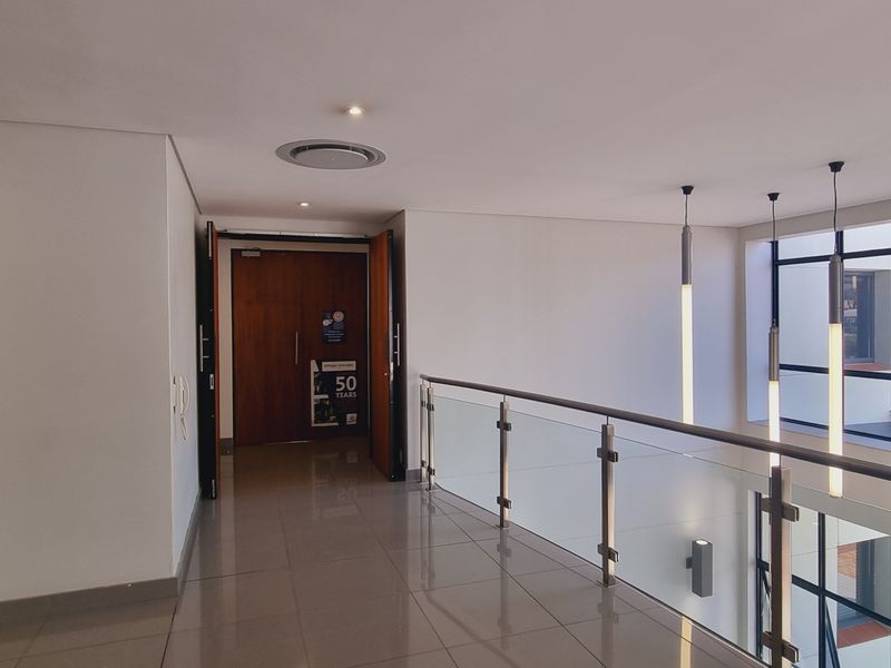 A bright and modern office hallway featuring polished tile floors, a glass balustrade, and contemporary pendant lighting with large windows overlooking the exterior.