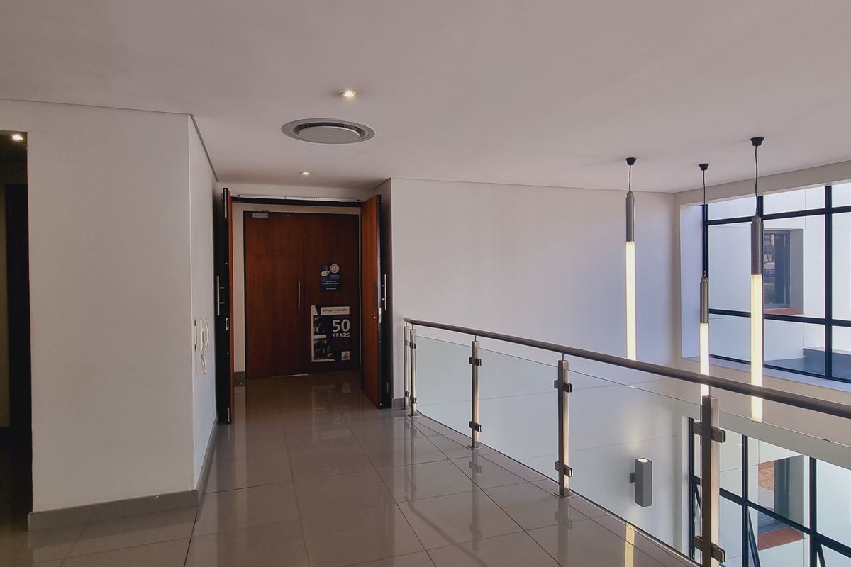 A bright and modern office hallway featuring polished tile floors, a glass balustrade, and contemporary pendant lighting with large windows overlooking the exterior.