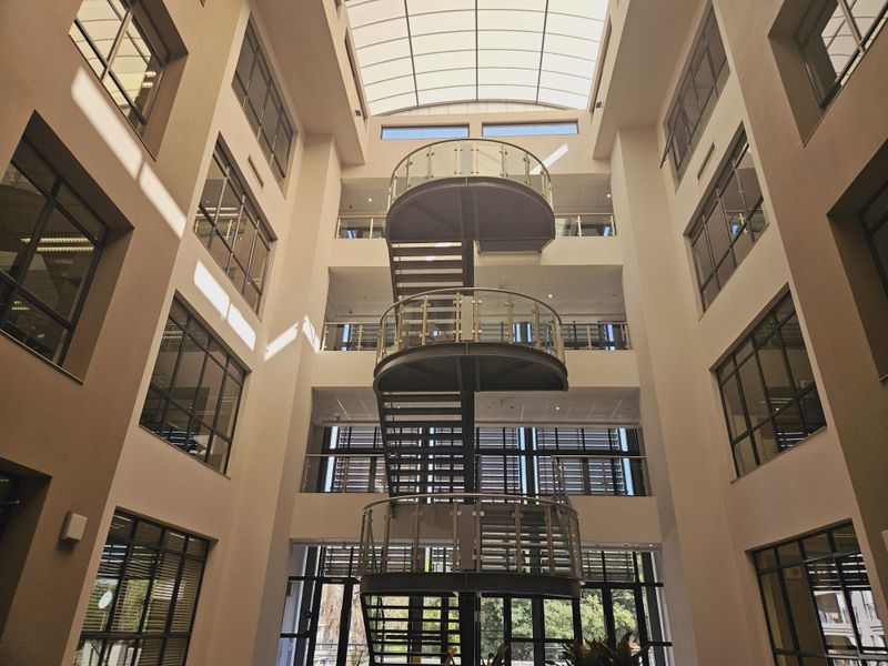 A bright, modern office atrium featuring a central spiral staircase, glass balustrades, and a large overhead skylight providing natural light.
