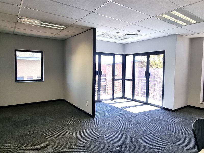 A bright, open-plan office space featuring carpet tiles, suspended ceilings with recessed lighting, and large glass doors leading to an exterior balcony.
