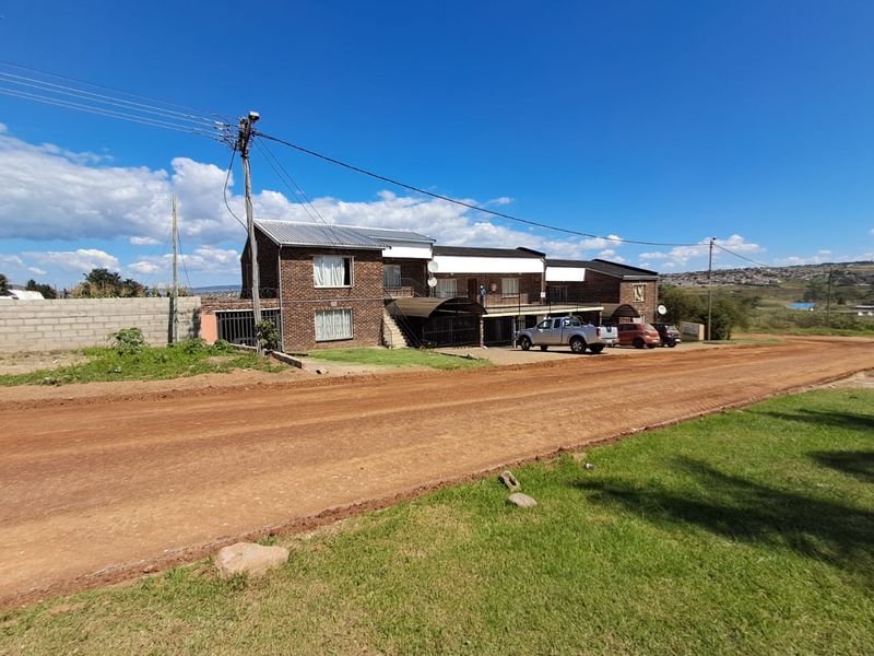 Exterior view of a face brick apartment building featuring a corrugated metal roof and secure gated access, situated along an unpaved road.