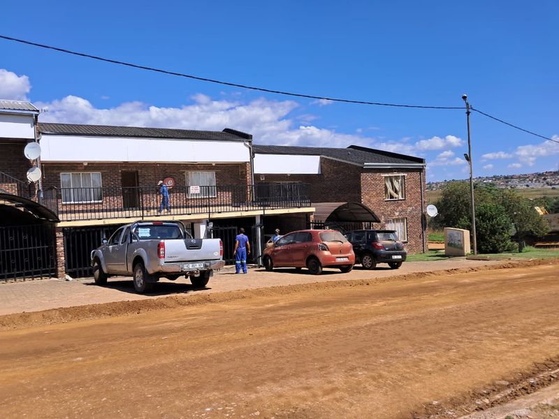 Front exterior view of a two-story face brick apartment complex featuring a balcony, paved parking area, and direct access from an unpaved road.