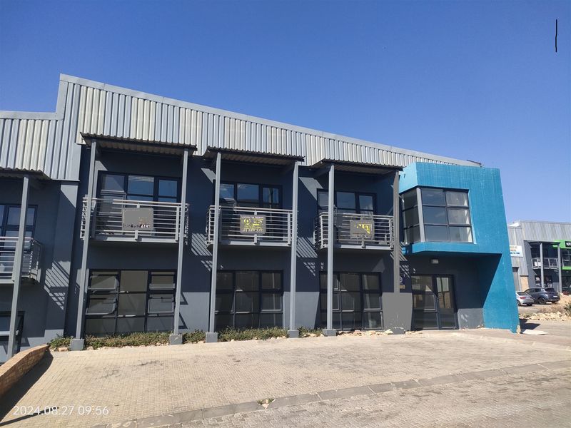 Modern industrial office or warehouse facade featuring grey metal cladding, blue accent wall, steel balconies, and large aluminium-framed windows with a paved frontage.