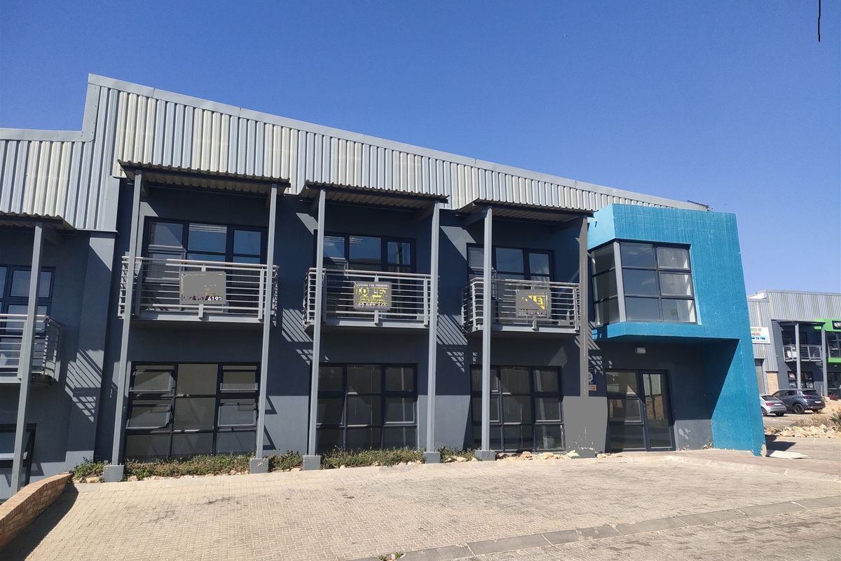 Modern industrial office or warehouse facade featuring grey metal cladding, blue accent wall, steel balconies, and large aluminium-framed windows with a paved frontage.