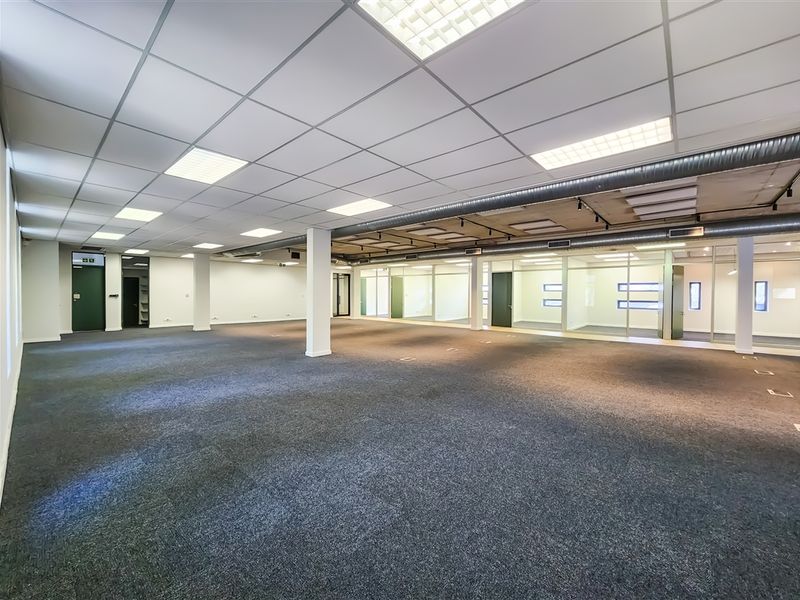Spacious open-plan office area featuring a suspended ceiling, recessed lighting, exposed industrial ducting, and glass-partitioned meeting rooms.
