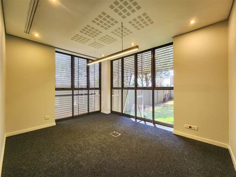 A bright, modern office space featuring large windows, carpeted flooring, integrated ceiling lighting, and a floor-mounted power outlet.