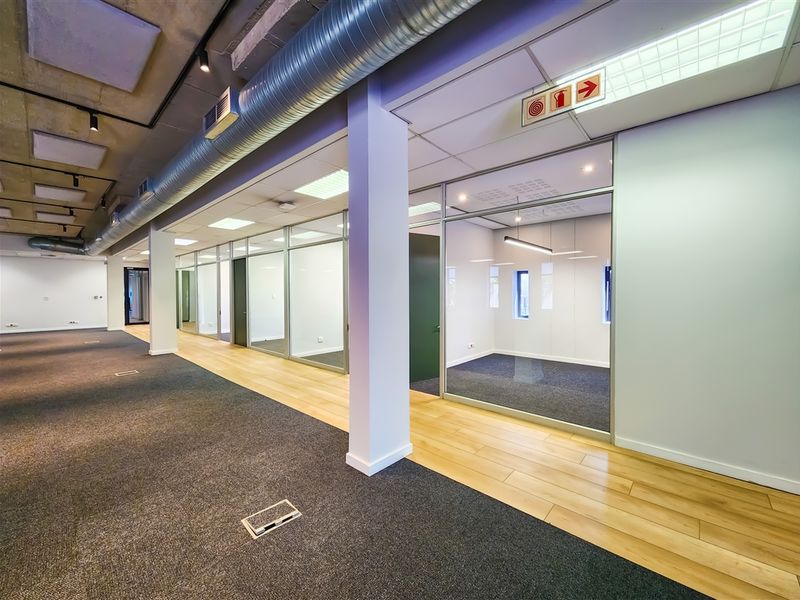 Modern open-plan office space featuring glass-partitioned private offices, exposed industrial ducting, and a mix of carpet and laminate flooring.