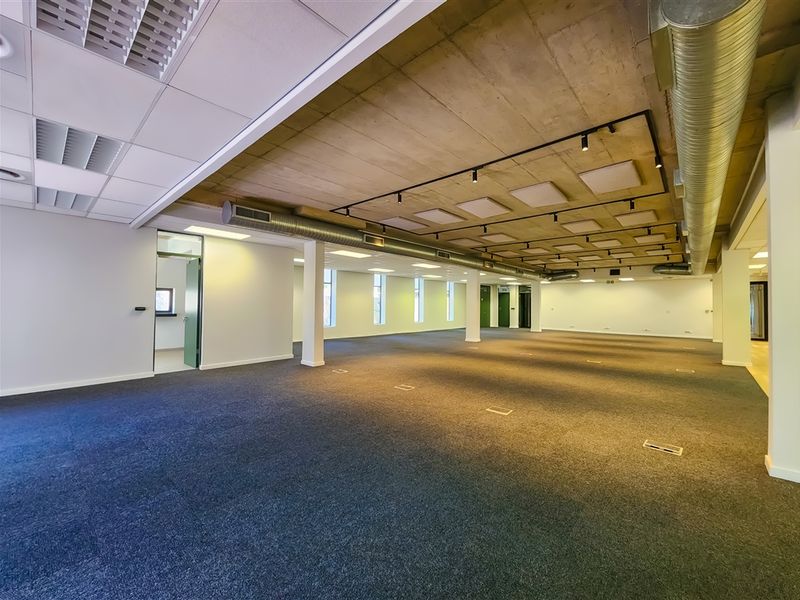 Spacious open-plan office area featuring an industrial-style exposed concrete ceiling, exposed ductwork, track lighting, and carpeted flooring with integrated floor power boxes.