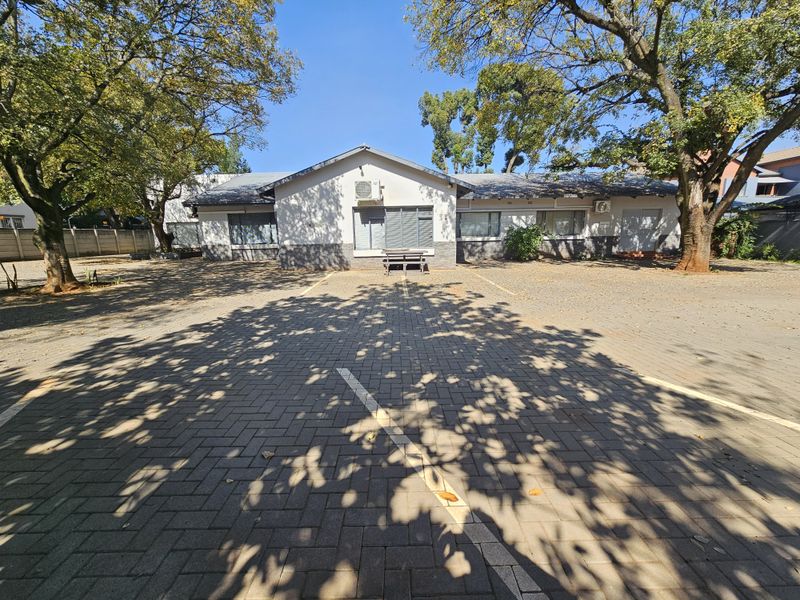Front elevation of the office building featuring a paved parking area, mature trees, and exterior security shutters on the windows.
