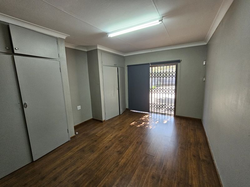 A clean, empty office space featuring laminate flooring, built-in storage cupboards, and a secure exterior access door with a security gate.