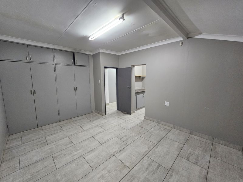 An empty office space featuring grey built-in cupboards, tiled flooring, and fluorescent lighting, with an adjacent small kitchenette area.