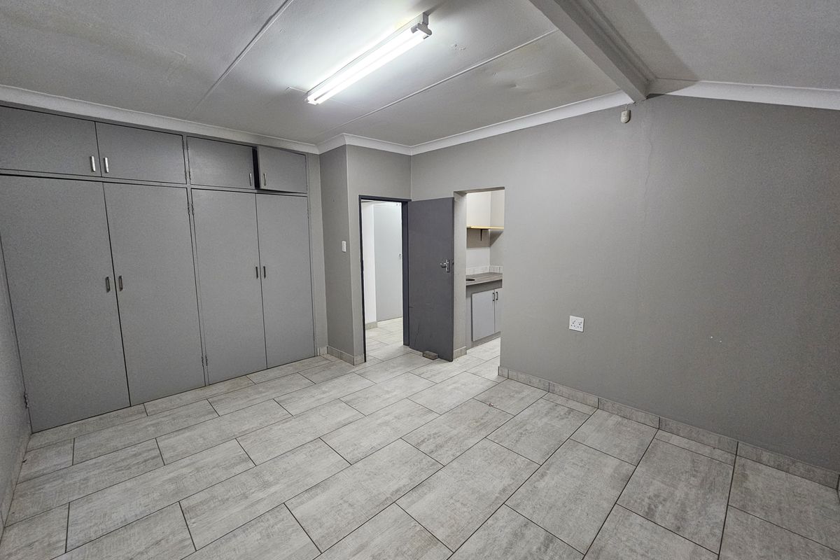 An empty office space featuring grey built-in cupboards, tiled flooring, and fluorescent lighting, with an adjacent small kitchenette area.