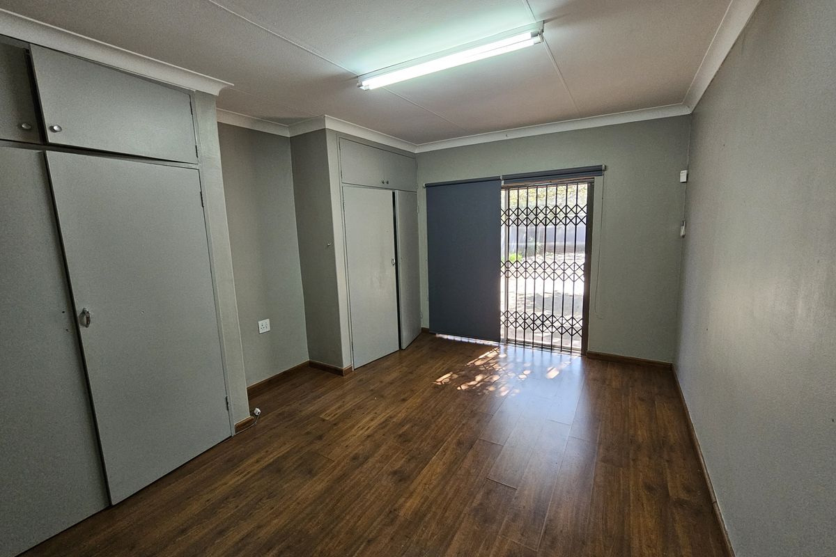 A clean, empty office space featuring laminate flooring, built-in storage cupboards, and a secure exterior access door with a security gate.