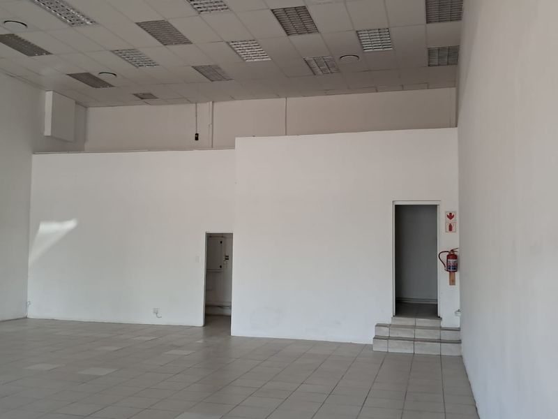 A spacious, empty retail unit featuring tiled flooring, a suspended ceiling with recessed lighting, and two internal access points.