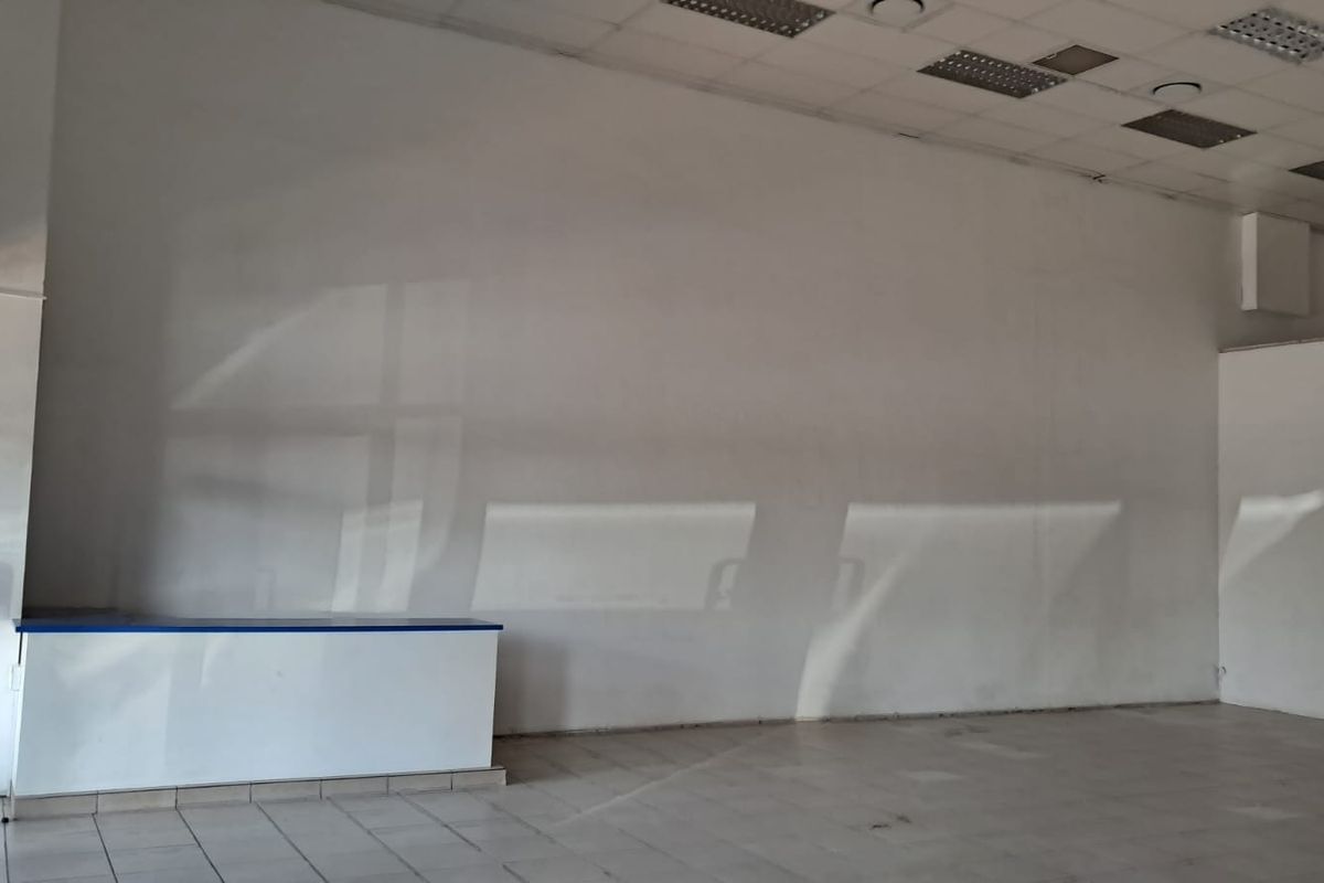 An empty, open-plan retail space featuring tiled flooring, a suspended ceiling with recessed lighting, and a built-in service counter.