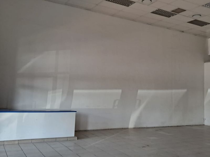 An empty, open-plan retail space featuring tiled flooring, a suspended ceiling with recessed lighting, and a built-in service counter.