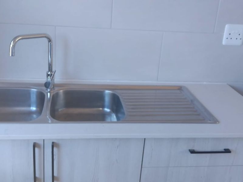 A clean, modern kitchen area featuring a stainless steel double sink, chrome mixer tap, light wood cabinetry, and a neutral tiled backsplash.