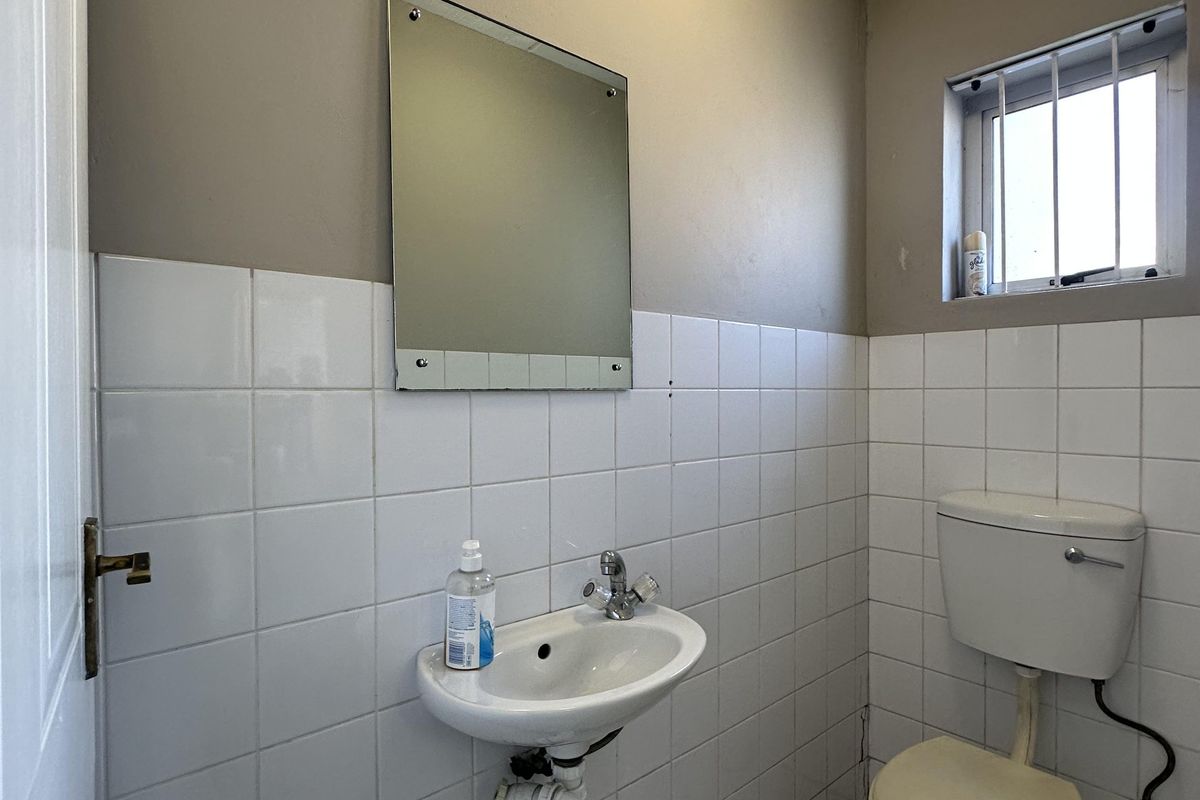 A compact industrial-style restroom featuring a wall-mounted basin, a standard toilet, and white wall tiling with a small security-barred window.