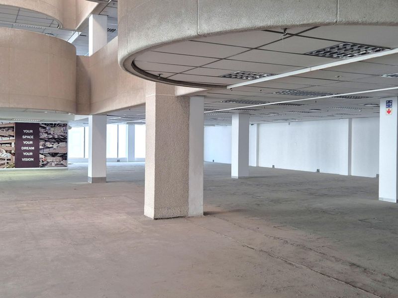 A large, open-plan office space featuring concrete structural columns, suspended ceilings with recessed lighting, and expansive windows providing natural light.