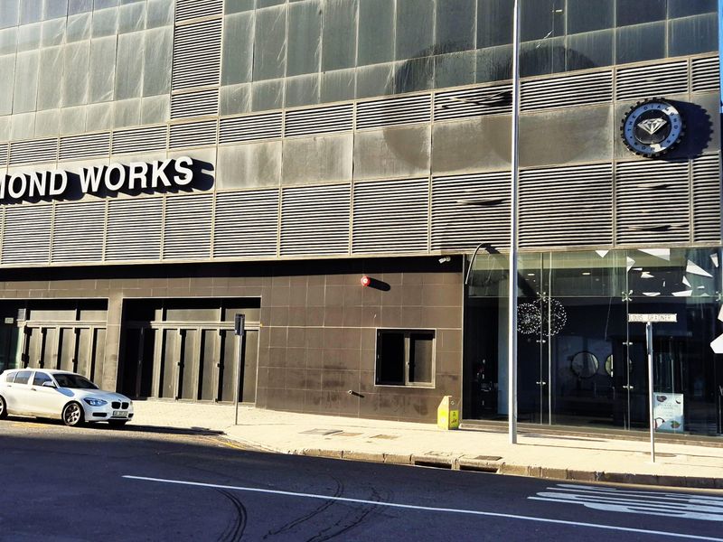 Front exterior view of a modern commercial office building featuring dark tiled cladding, metal louvers, and large glass entrance panels.