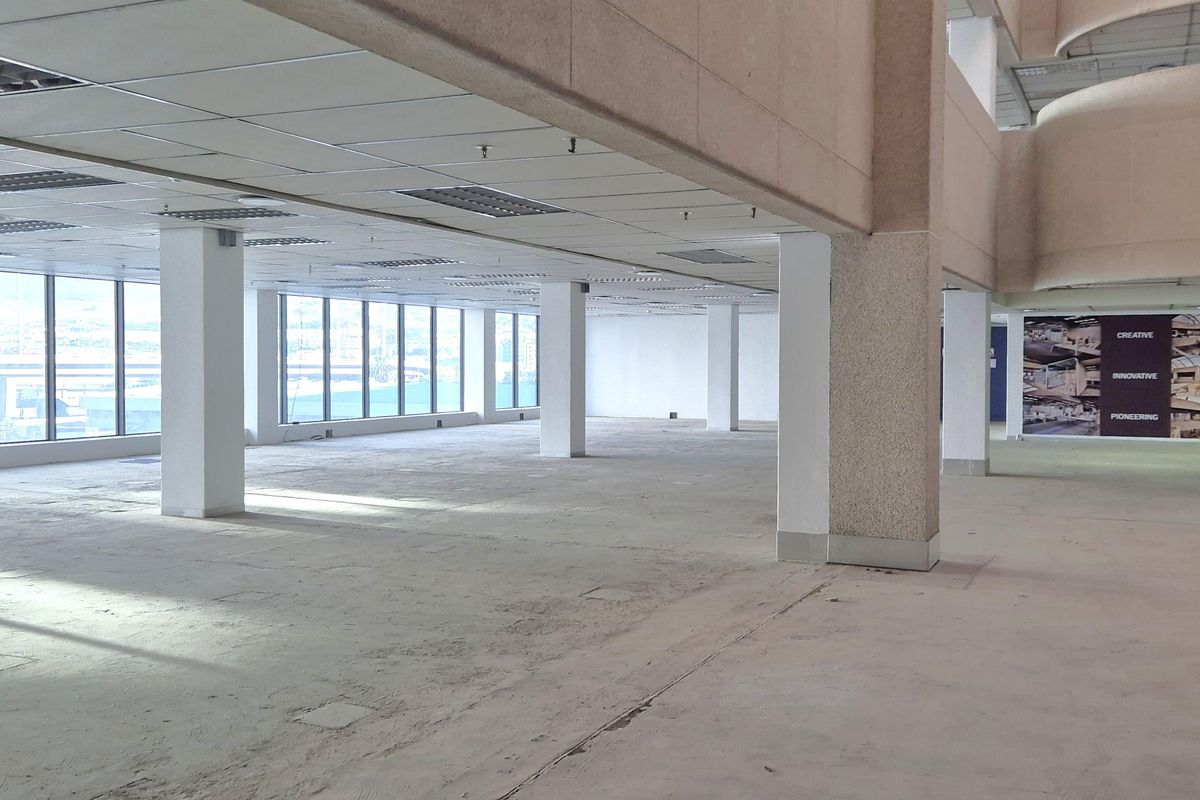 A large, open-plan office space featuring expansive windows, structural columns, and a suspended ceiling, currently in a vacant, shell-like condition.