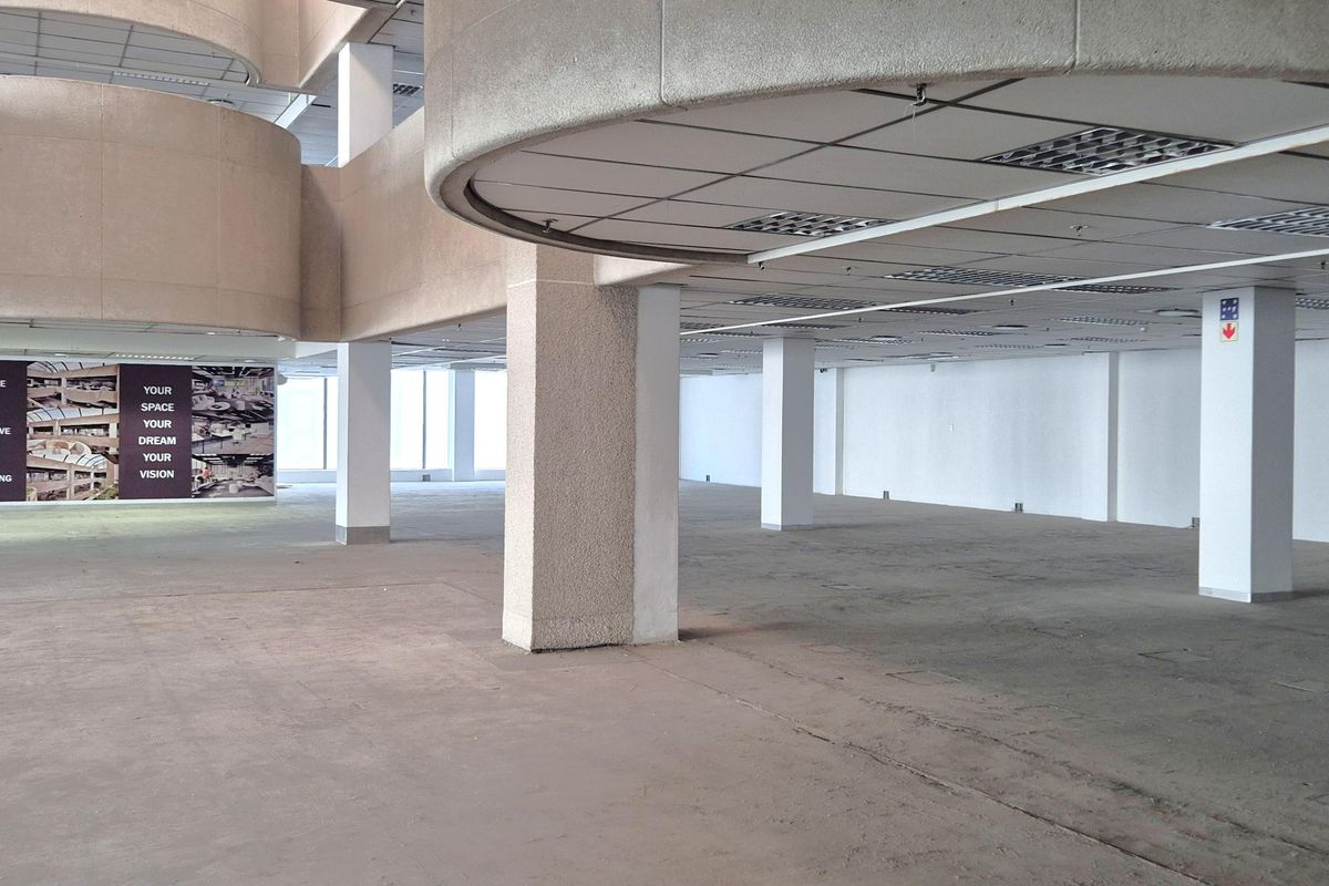 A large, open-plan office space featuring concrete structural columns, suspended ceilings with recessed lighting, and expansive windows providing natural light.