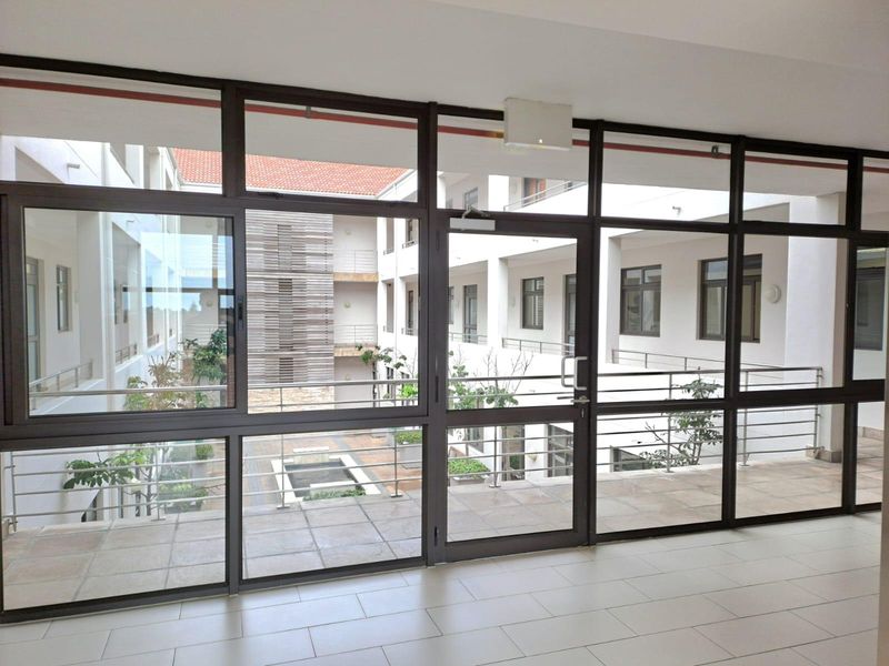 A bright, modern office hallway featuring extensive aluminium-framed glass partitioning and a glass door, overlooking an internal courtyard with tiled flooring.