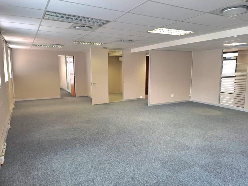 A spacious, open-plan office area featuring neutral walls, carpet tile flooring, and a suspended ceiling with integrated lighting.