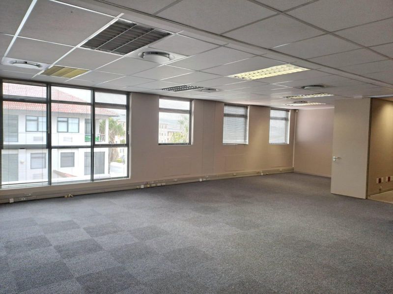 A spacious, empty office area featuring carpet tiles, a suspended ceiling with recessed lighting, and large windows with venetian blinds.