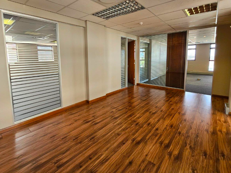 A bright, vacant office space featuring wood laminate flooring, glass partitions, and a suspended ceiling with integrated lighting.