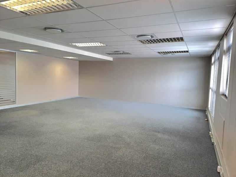 A spacious, empty open-plan office area featuring a suspended ceiling with recessed lighting, fitted carpeting, and large windows with venetian blinds.