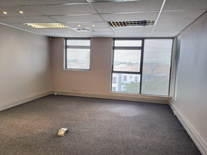 A vacant, well-lit office space featuring a suspended ceiling, carpeted flooring, and large windows with venetian blinds, offering a functional professional environment.