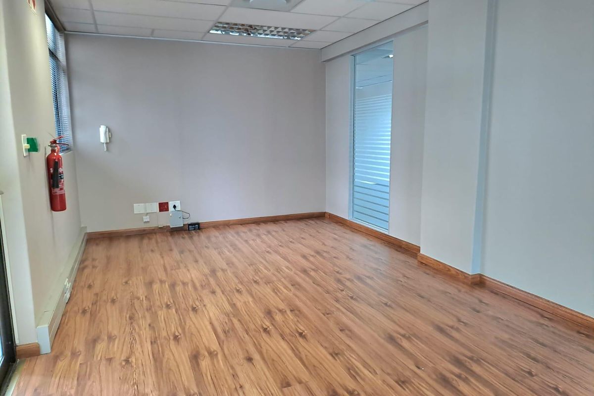 A clean, empty office space featuring laminate wood flooring, a suspended ceiling with recessed lighting, and a glass partition wall.