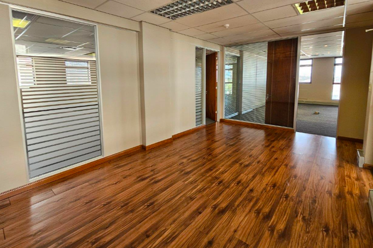 A bright, vacant office space featuring wood laminate flooring, glass partitions, and a suspended ceiling with integrated lighting.
