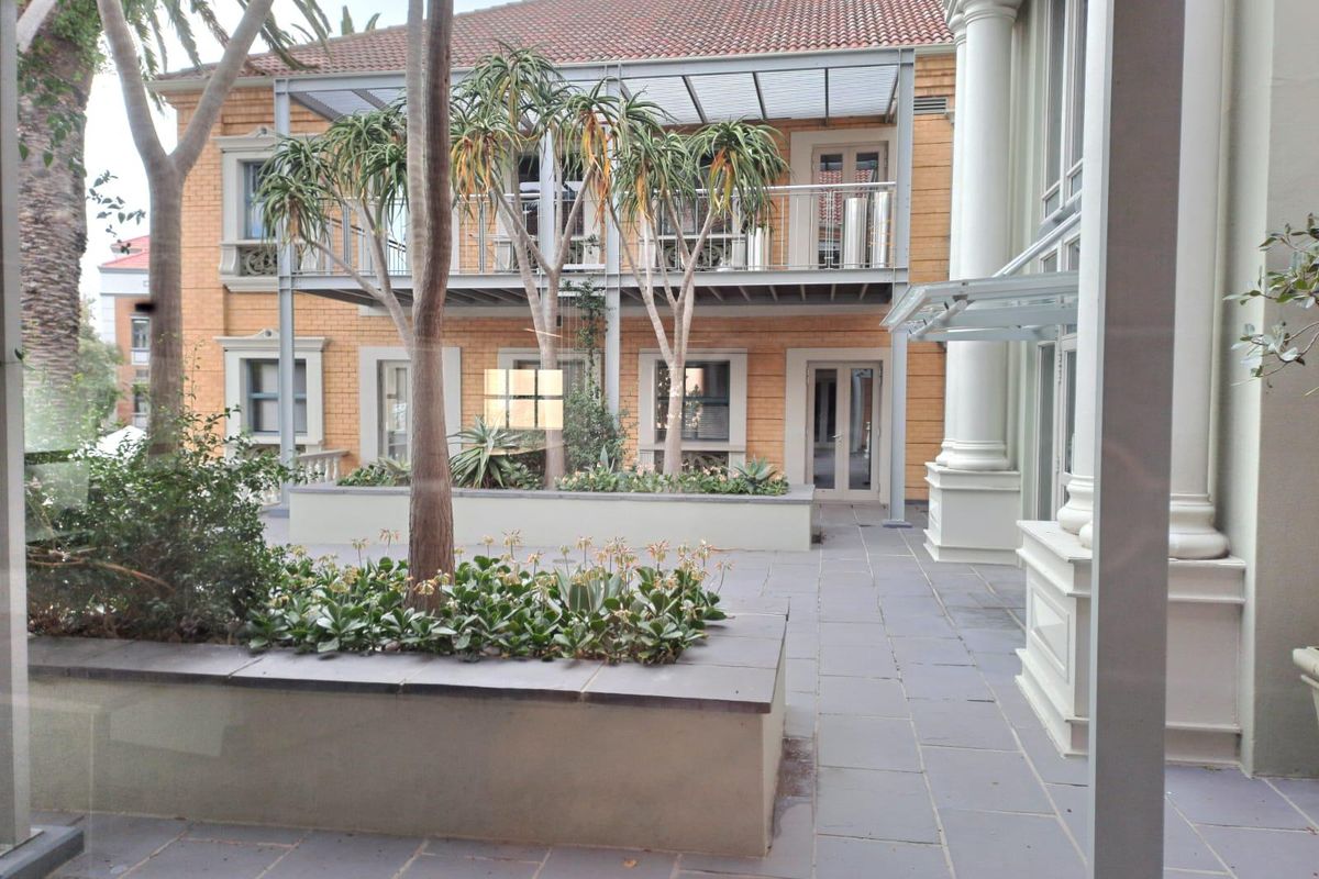 A view of a well-maintained courtyard featuring stone paving, raised garden beds, and a multi-story building with a balcony and classic architectural columns.
