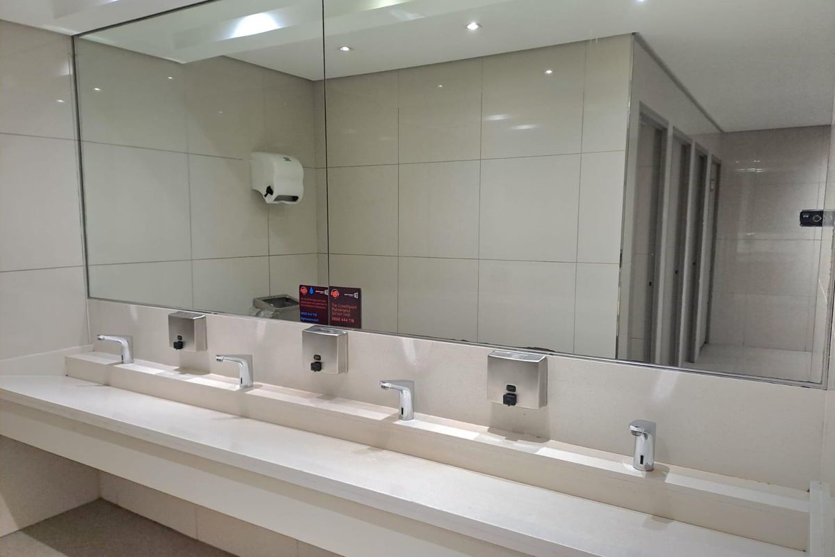 Modern commercial office restroom featuring a long vanity with multiple integrated sensor faucets, soap dispensers, and a large wall-to-wall mirror.
