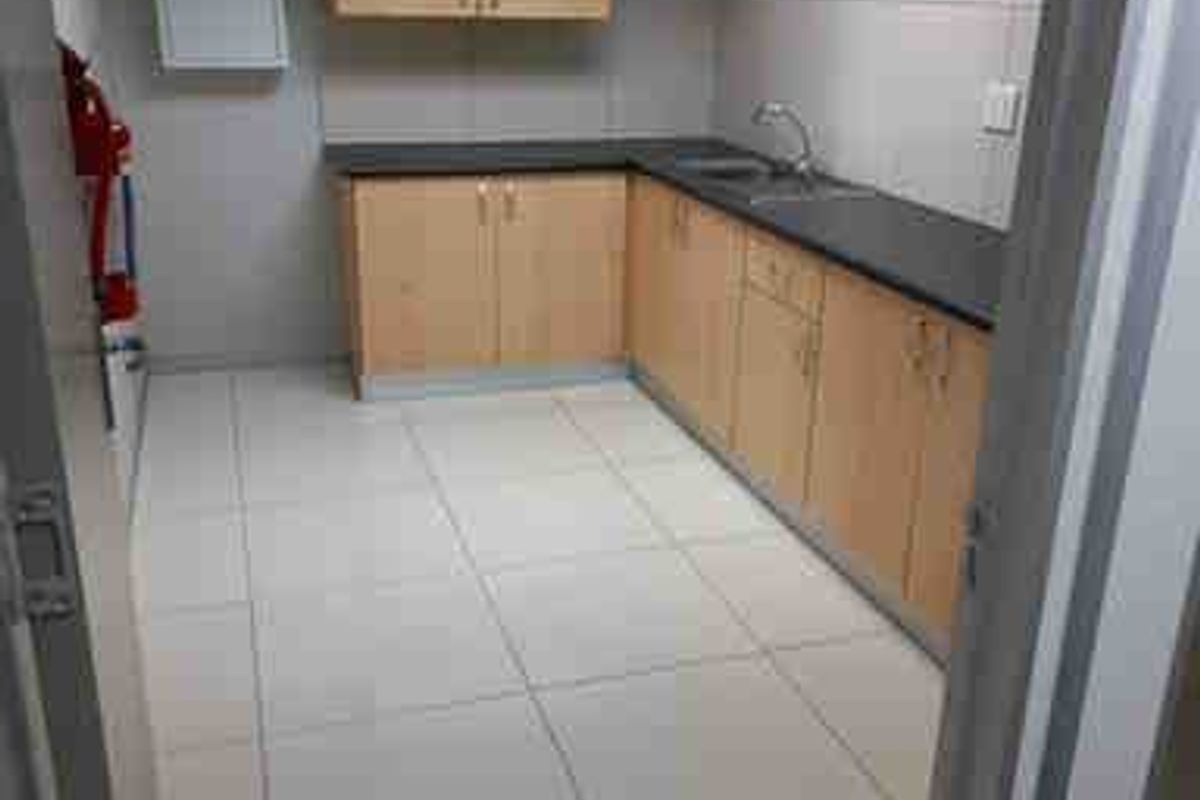 A clean, functional commercial kitchenette featuring light wood-finish cabinetry, dark countertops, a stainless steel sink, and bright tiled flooring.