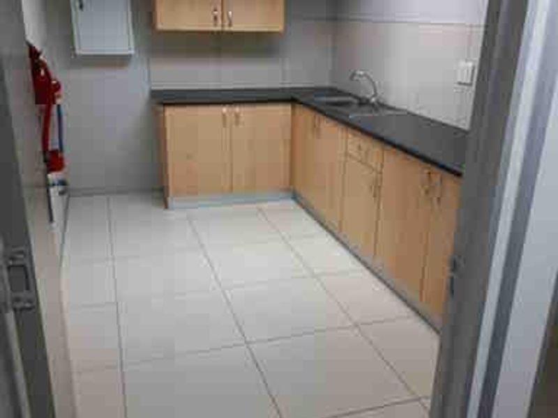 A clean, functional commercial kitchenette featuring light wood-finish cabinetry, dark countertops, a stainless steel sink, and bright tiled flooring.