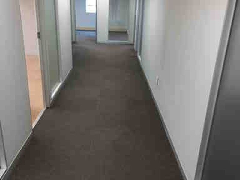 A functional commercial office hallway featuring carpeted flooring, suspended ceilings with recessed lighting, and glass partition doors leading to adjacent office spaces.