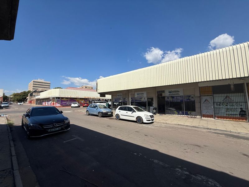 Street-level view of a commercial retail strip featuring shopfronts with security gates and a corrugated metal roof structure.