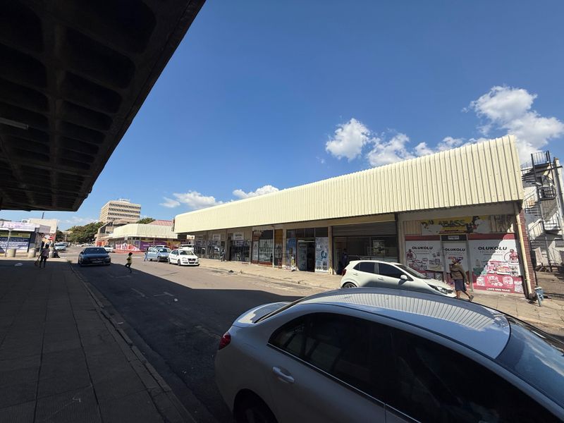 Street-level view of a commercial strip with multiple storefronts, paved sidewalk, and metal roofing under a clear sky.