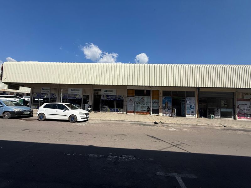 Front elevation of a commercial strip mall featuring multiple storefronts with security gates and a large corrugated metal canopy.
