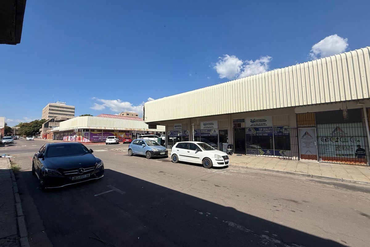 Street-level view of a commercial retail strip featuring shopfronts with security gates and a corrugated metal roof structure.