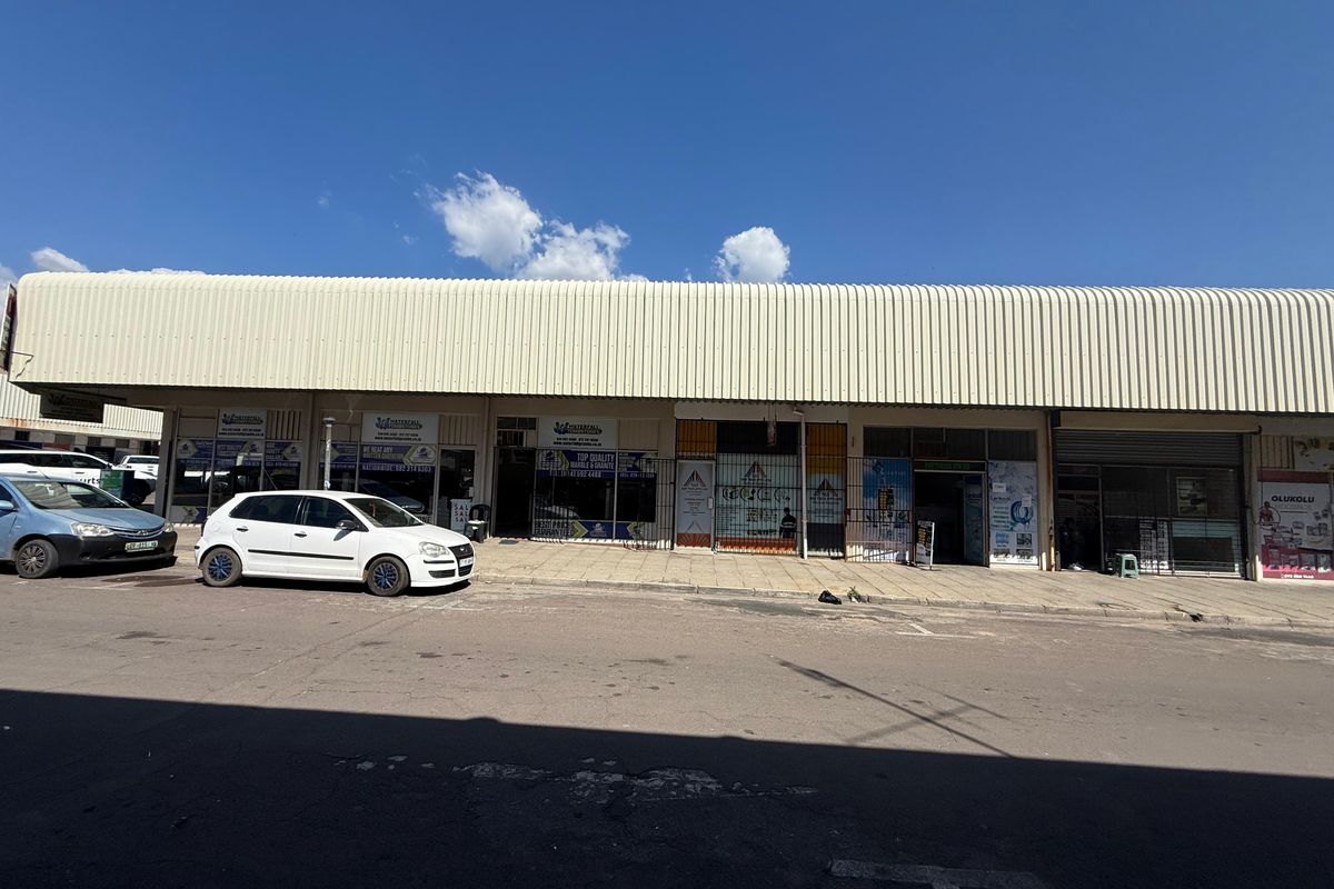 Front elevation of a commercial strip mall featuring multiple storefronts with security gates and a large corrugated metal canopy.