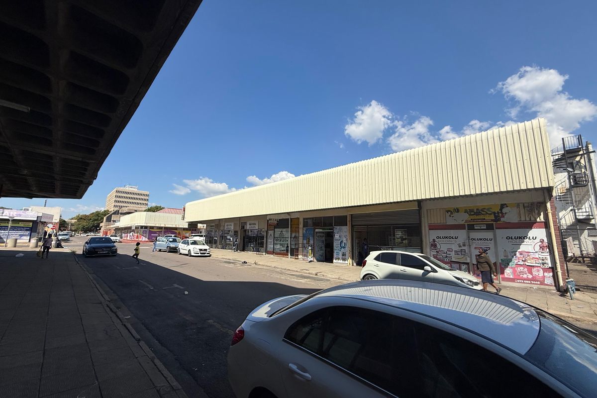 Street-level view of a commercial strip with multiple storefronts, paved sidewalk, and metal roofing under a clear sky.