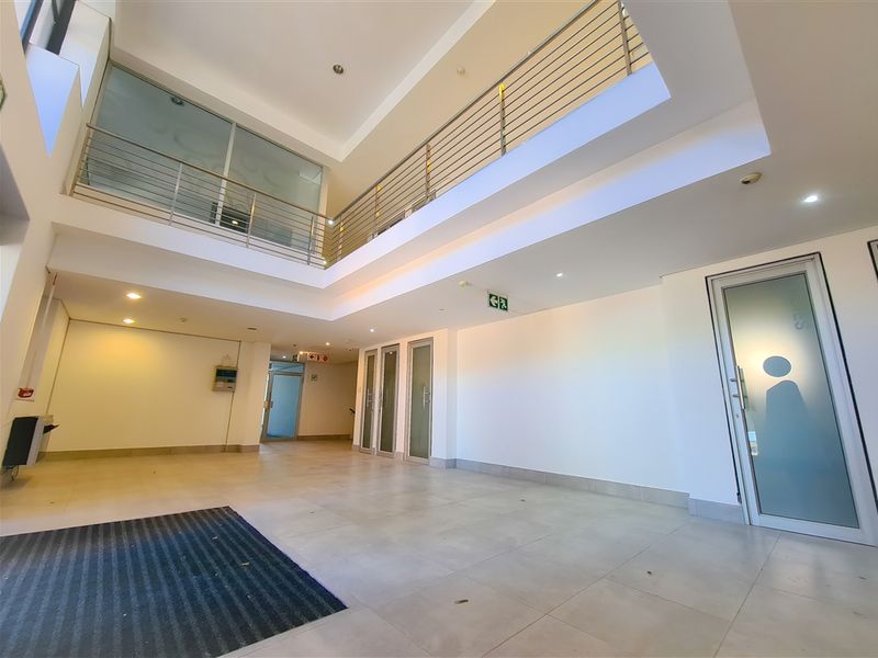 Spacious double-volume office building entrance lobby featuring tiled flooring, a mezzanine level with stainless steel balustrades, and modern glass-panelled doors.