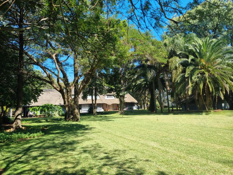A spacious, well-maintained garden featuring mature trees and a view of a traditional thatched-roof farm dwelling.