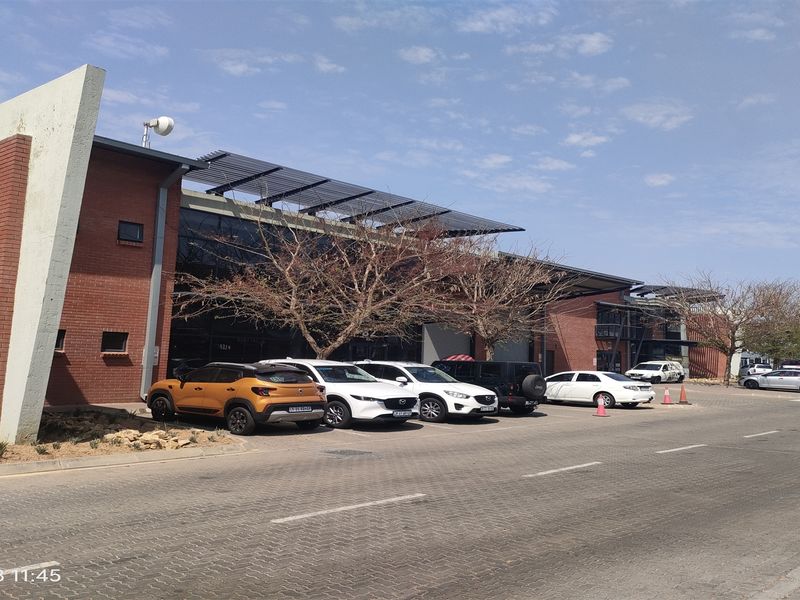 Front elevation of a modern industrial building featuring exposed brickwork, large glass facade, and roof-mounted solar panels with an adjacent paved parking area.