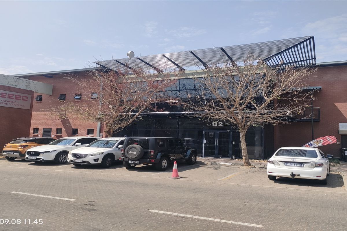 Front exterior view of an industrial office building featuring a brick facade, glass curtain wall entrance, and dedicated paved parking area.